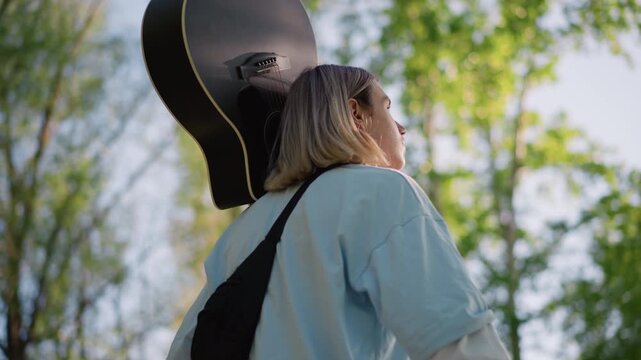 young white guitarist carrying guitar case through leafy park, head tilted upward toward sky, confident busker energy and touring songwriter vibe, sundappled background, candid handheld shots, upbeat