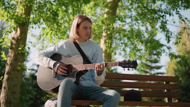 young white guitarist playing acoustic guitar on wooden bench under birch trees, casual jeans and sweater, focused expression, fingerpicking and strumming, open guitar case beside, warm afternoon