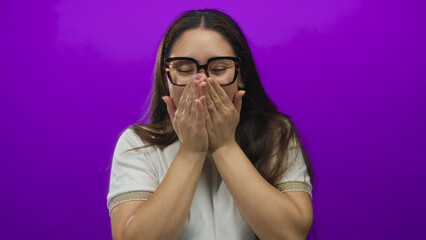 Fototapeta premium Young hispanic woman wearing glasses raises palms and covers mouth in purple studio; celebration joy.