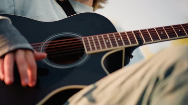 young person playing black acoustic guitar outdoors, unspecified musician gazing into distance, soft sunset light, casual sweater, finger on strings, intimate portrait with cinematic bokeh and relaxed