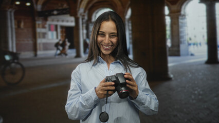 Young hispanic woman holding dslr camera and smiling in a city street archway while checking photos on the camera screen  joy creativity travel adventure. © Krakenimages.com