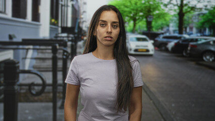 Fototapeta premium Woman standing on street in a city, visible face and long hair, neutral expression and relaxed posture with arms at sides; contemplation.