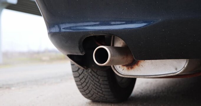 Close-up view of a car exhaust pipe showing gradual angle change, revealing tire tread and rust details, set against an outdoor backdrop with a blurred structure