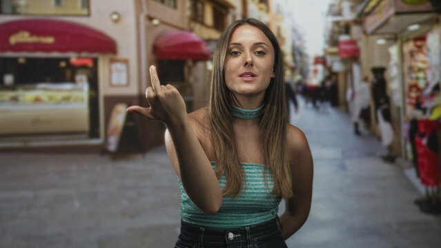Woman shows middle finger toward camera on a narrow pedestrian street with shop awnings and blurred passersby; defiance.