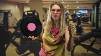 Fototapeta premium Blonde woman holding vinyl record, fist clenched and singing while wearing pink sunglasses in a gym building; retro workout fun.