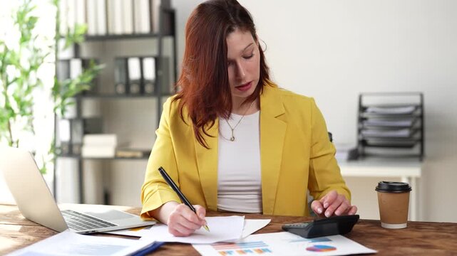 Professional businesswoman using a calculator and writing notes on financial documents with charts, then cross-referencing information on her laptop computer while working in a modern office
