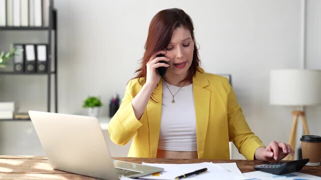 Frustrated female accountant having a difficult conversation on her smartphone while calculating finances and using a laptop at her desk, showing clear signs of stress and anger