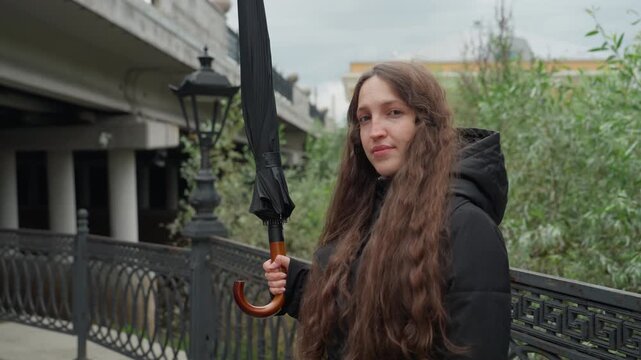 young woman holding umbrella on bridge, city walkway with iron railing and lampposts, overcast sky, long curly hair spilling over black parka hood, gentle smile pausing during walk, autumn foliage