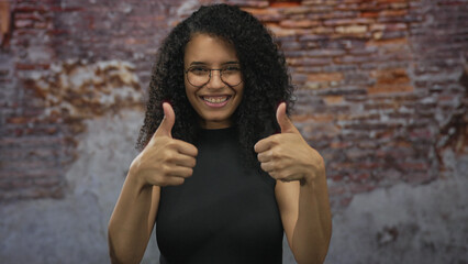 Fototapeta premium Young hispanic woman with glasses smiling shows thumbs up over isolated brick building wall; approval.