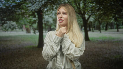 Obraz premium Woman clutching neck with hand, striped shirt visible, worried expression in forest with trees and grass; anxiety solitude.