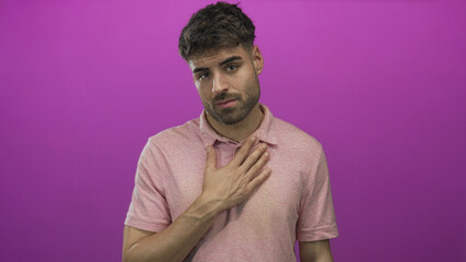 Young hispanic man with serious expression hold hand on chest over pink polo in purple studio...