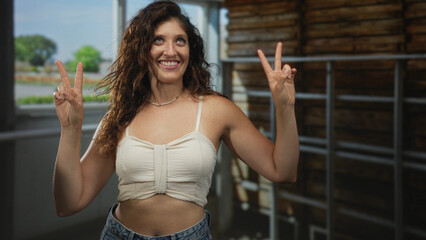 Fototapeta premium Woman showing double peace signs and bare midriff in studio space with window light and wooden rack; joy celebration.