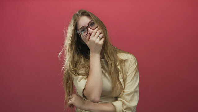 Young blonde woman with long hair and glasses covers mouth with hand while tilting head in studio; amusement.