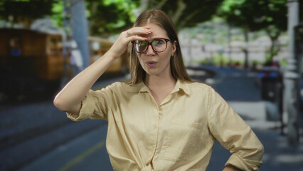 Fototapeta premium Woman wearing glasses and beige shirt touching forehead with hand on street under green trees; confusion.