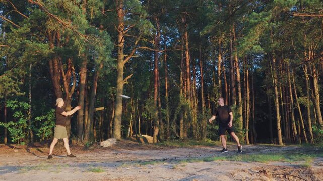 Two men playing frisbee in forest sandy clearing beside tall pine trunks warm golden light dynamic tosses catches casual shorts sneakers energetic friendship outdoor recreation at sunset