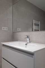 Fototapeta premium Modern bathroom interior with a sleek white sink, chrome faucet, and gray tiled walls. A mirror reflects the minimalist design and a towel radiator is visible.