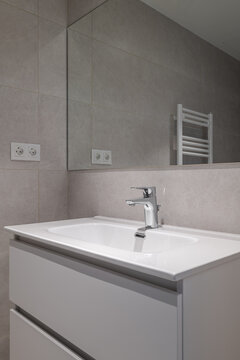 Modern bathroom interior with a sleek white sink, chrome faucet, and gray tiled walls. A mirror reflects the minimalist design and a towel radiator is visible.