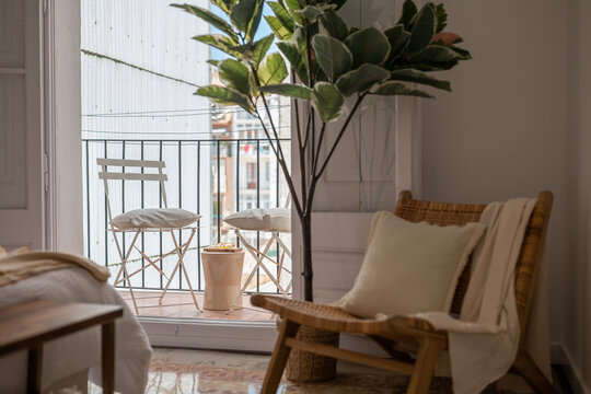 A cozy interior scene featuring a wooden chair, a potted plant, and a balcony view. Natural light illuminates the space, creating a warm atmosphere.