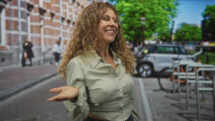 Fototapeta premium Woman hand to lips blowing a kiss, smiling and gesturing with open palm beside a parked car and bike rack on a city street with brick building and trees; joyful warmth.