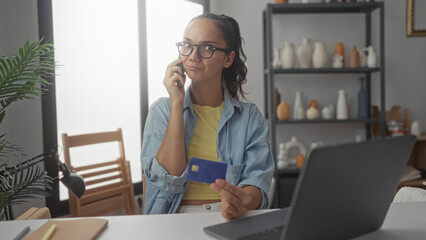 Young hispanic woman phone to ear holding credit card by laptop in a studio with skeptical pout...
