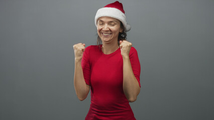 Fototapeta premium Woman in red shirt and santa hat clenches fists, raises arms and smiles in studio; holiday excitement.