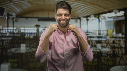 Fototapeta premium Hispanic man with beard raises clenched fists and smiles amid empty cafe tables under arched ceiling in a building; joy celebration.