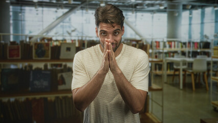 Naklejka premium Man with hands pressed together in a pleading gesture inside a building library with visible bookshelves; anxiety reflection.