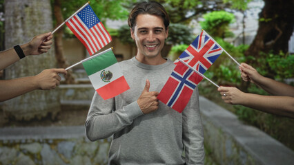 Fototapeta premium Man holding three small national flags from usa mexico norway and giving thumbs up in forest pathway, smiling broadly; unity celebration joy.