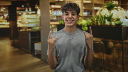 Obraz premium Teen boy in gray tshirt makes rock horns with both hands, smiles broadly and shows a silver chain in a cafe building; playful joy.