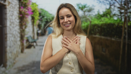 Naklejka premium Woman blonde smiling with hands crossed on chest on a street with stone wall and greenery; gratitude connection.
