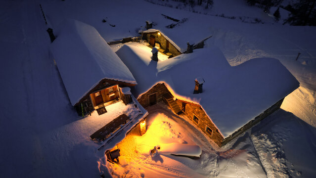 Aerial view of snow-laden chalets glow warmly against the frigid, dark landscape, casting a golden hue on the pristine white snow, Valgrisenche, Aosta Valley, Italy.