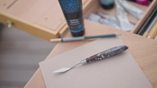 palette knife rests on paper next to paint tube and pencil, closeup on textured surface, studio prep for impasto techniques, metallic blade with paint residue, soft shallow depth, concentrated mood,