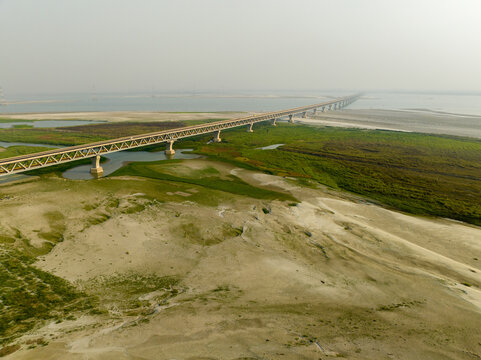 Aerial view of the Padma Multipurpose Bridge stretches across the mighty river, connecting vibrant green landscapes with sandy shores, Mawa, Dhaka Division, Bangladesh.