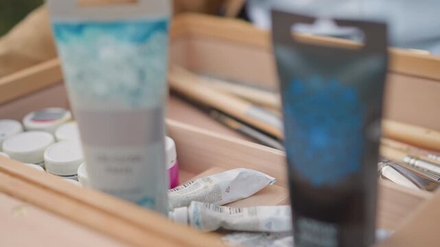 paint tubes brushes on wooden easel, plein air street artist preparing palette and canvas, closeup of jars and brushes inside wooden box, textured paper for sketch, soft natural light, neutral color