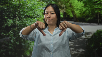 Naklejka premium Woman showing thumbs down gesture with bare hands in park among green trees and a paved path; disapproval criticism.
