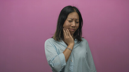 Fototapeta premium Woman touching cheek and jaw with hand in pink studio wearing light blue shirt and bracelet, slight frown and hand at jawline; toothache concern.