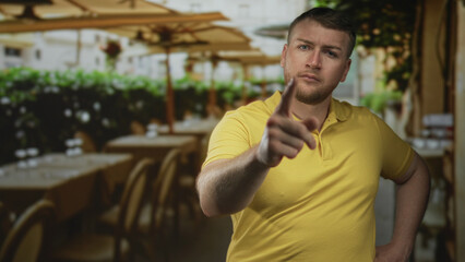 Obraz premium Young overweight man in yellow polo shirt points finger at camera on street terrace restaurant outdoor seating; anger confrontation.