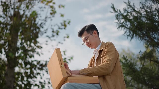 man in park using wooden laptop, sketching and typing on vintage case while sunlight filters through trees, casual jacket and bench seating, relaxed focused mood, writer, illustrator and student roles
