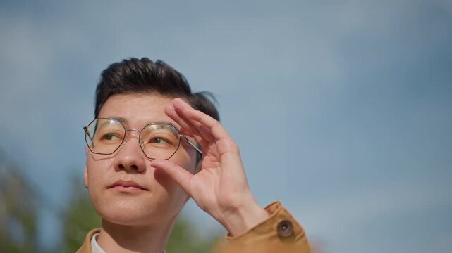 Asian illustrator refining portrait on easel, adjusting glasses while studying composition midshot and profile frames reveal deliberate brushwork, calm mood, leafy backdrop, soft light, cinematic