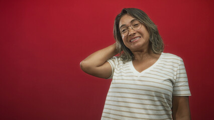 Fototapeta premium Woman touches neck with hand in red studio, wearing striped t shirt and glasses showing a wry grimace; mild embarrassment.