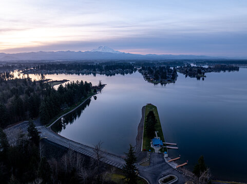 Aerial view of the serene lake reflecting the pastel sky, framed by dark, dense trees and distant, snow-capped mountains, Lake Tapps, Washington, United States.