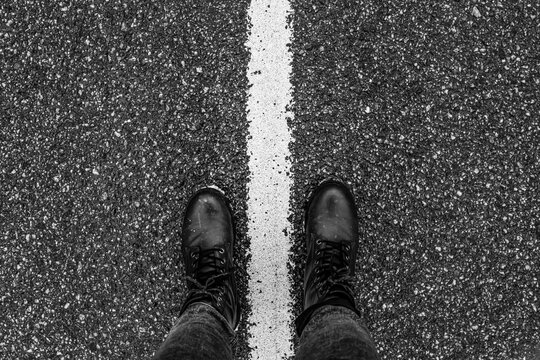 Top view of black leather boots standing on a white road marking over textured asphalt surface. Urban minimal composition with strong contrast and centered alignment