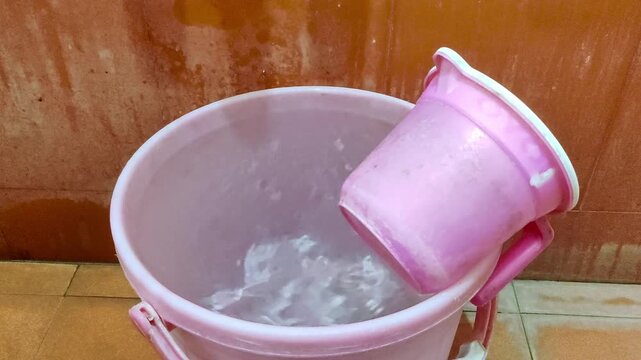 A close-up of a pink plastic water bucket filled with water, with a bathing mug resting beside it.