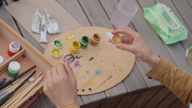 hands mixing acrylic paint on palette while preparing outdoor sketching setup with brushes, jars, wipes and wooden table focused creative process with vivid pigments and textured strokes