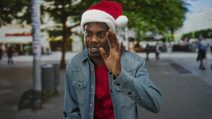 Fototapeta premium Man wearing santa hat and denim jacket waves hand with friendly smile on lively city street lined with trees; holiday joy.