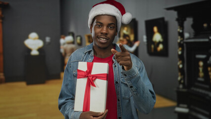 Fototapeta premium Man wearing santa hat and denim jacket holding white gift box wrapped in red ribbon and pointing finger in building; holiday joy.