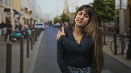Fototapeta premium Woman clutching shoulder on a bustling city street near scooters and bollards, grimacing while wearing a gold necklace and denim jeans; discomfort vulnerability.