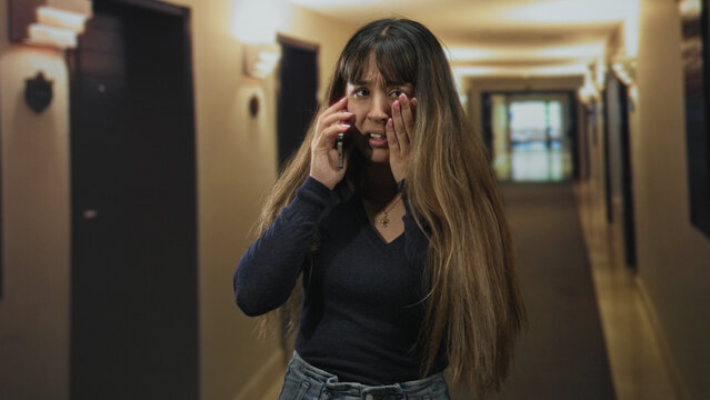 Young hispanic woman on phone wiping tears with hand and covering face in dim hotel hallway; sadness vulnerability.