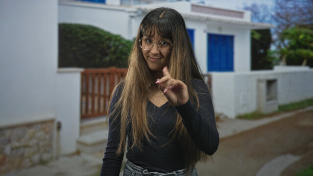 Woman wearing glasses and necklace points finger with a casual stance on street near a white building and blue door, long hair framing face; playful confidence.