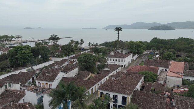 Red Colonial Rooftops Lead Toward the Palm-lined Waterfront and Calm Bay in Paraty, Brazil - Orbit Drone Shot